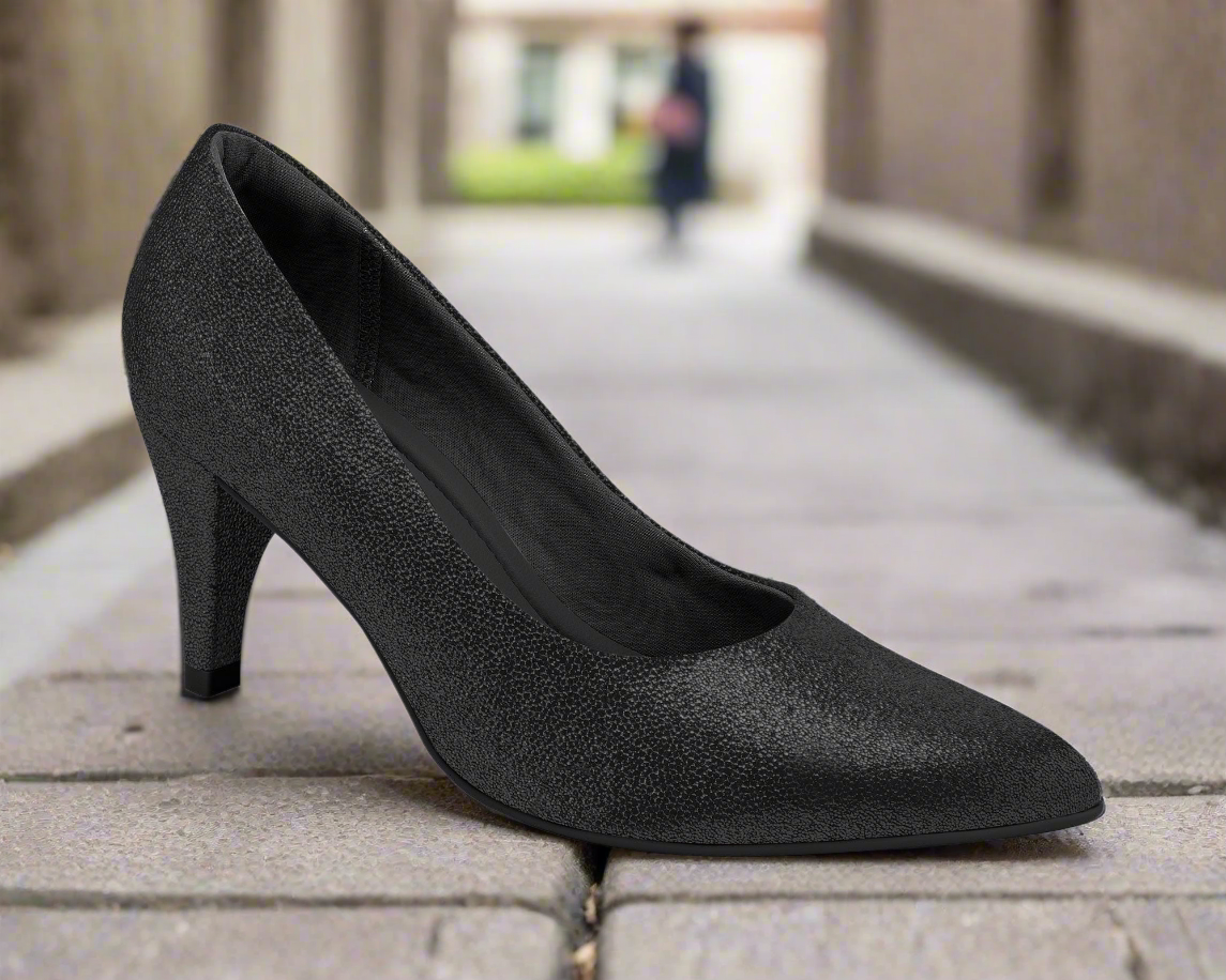 The Piccadilly Working Woman Heel - Black Glitter (Ref 745035-796) sits on a cobblestone path, with blurred buildings and a person in the background, showcasing a stylish yet comfortable choice for professional women.