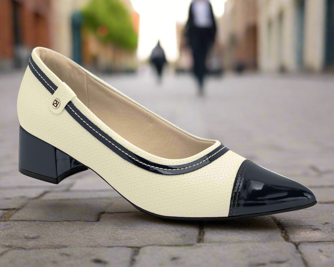 The Piccadilly Professional Comfort Shoe - Travel & Work (Ref 739052-01) in cream with a glossy black toe cap and trim is shown on a cobblestone street, with blurred figures and buildings in the background.