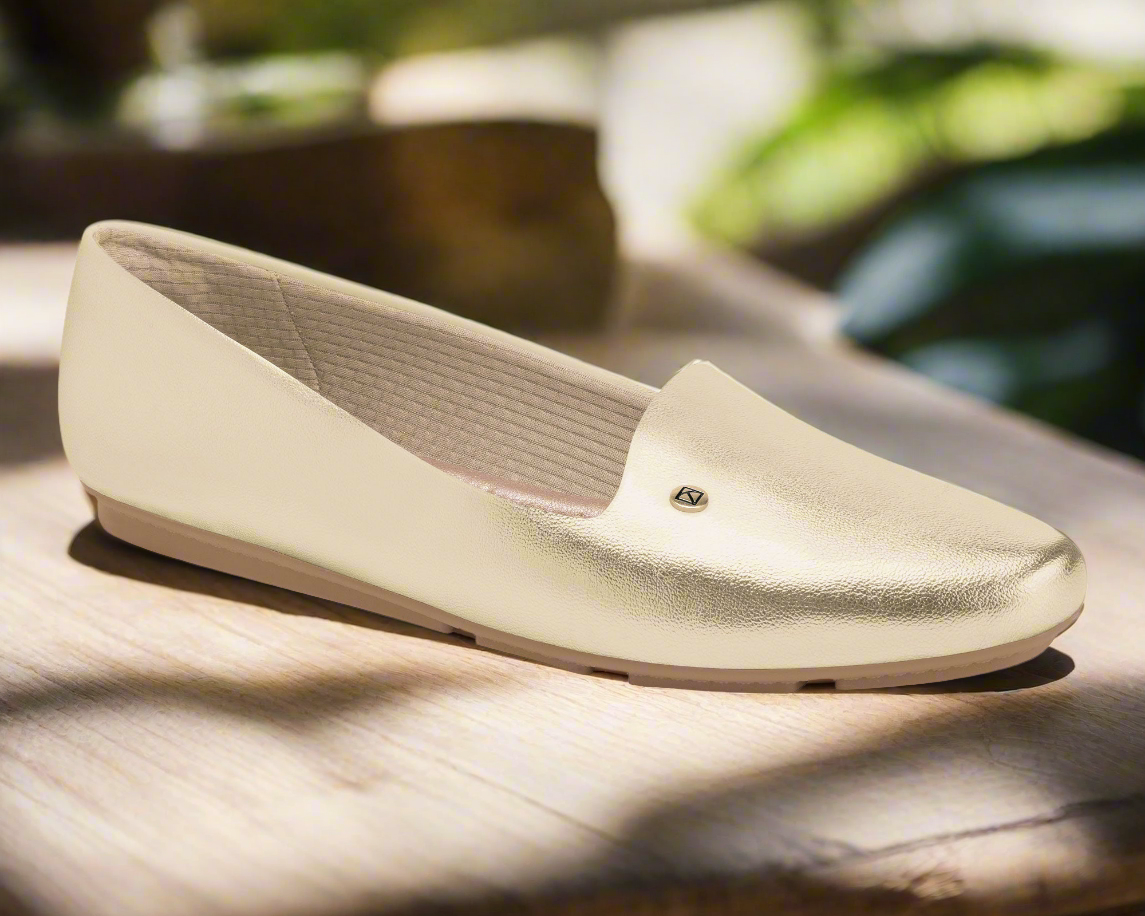A single Piccadilly Class & Comfort Moccasin Flat - Florence Gold (Ref 122007-12) with a metallic logo and cushioned insole rests on wood under soft sunlight, blending comfort and style for travel amid a serene, green backdrop.